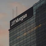 A glass skyscraper with the illuminated "J.P. Morgan" logo near the top, photographed at sunset with a cloudy sky in the background, highlights the firm’s influence on markets like Apple stock and its recent AAPL target announcements.