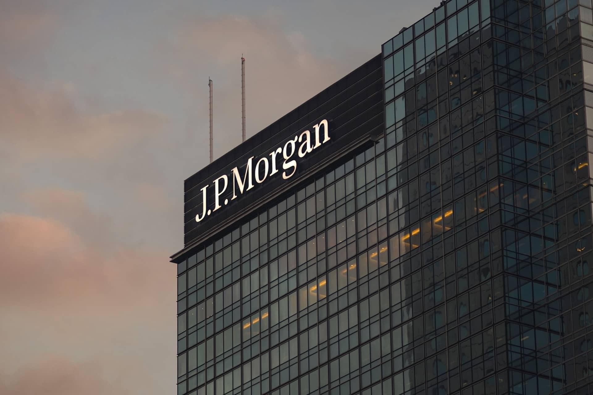 A glass skyscraper with the illuminated "J.P. Morgan" logo near the top, photographed at sunset with a cloudy sky in the background, highlights the firm’s influence on markets like Apple stock and its recent AAPL target announcements.