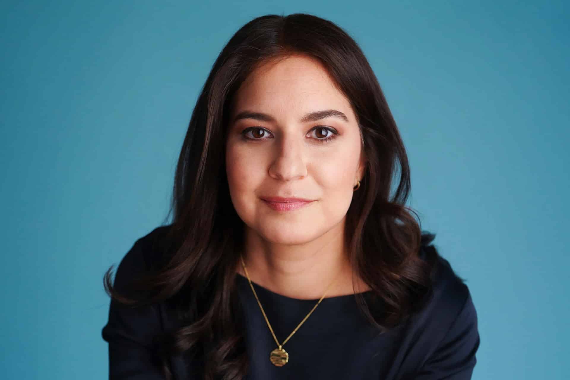 Woman with long dark hair wearing a navy top and gold necklace, looking at the camera while seated in front of a solid blue background—a striking image in light of recent discussions about AI-generated books and their growing impact on the publishing industry.