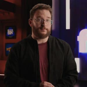 A man with short brown hair, glasses, and a beard stands indoors, wearing a maroon shirt and black jacket. Behind him are illuminated signs and a blurred blue logo—reminiscent of the backgrounds seen in Jon Prosser’s iOS 26 leaks videos.