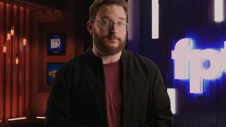 A man with short brown hair, glasses, and a beard stands indoors, wearing a maroon shirt and black jacket. Behind him are illuminated signs and a blurred blue logo—reminiscent of the backgrounds seen in Jon Prosser’s iOS 26 leaks videos.