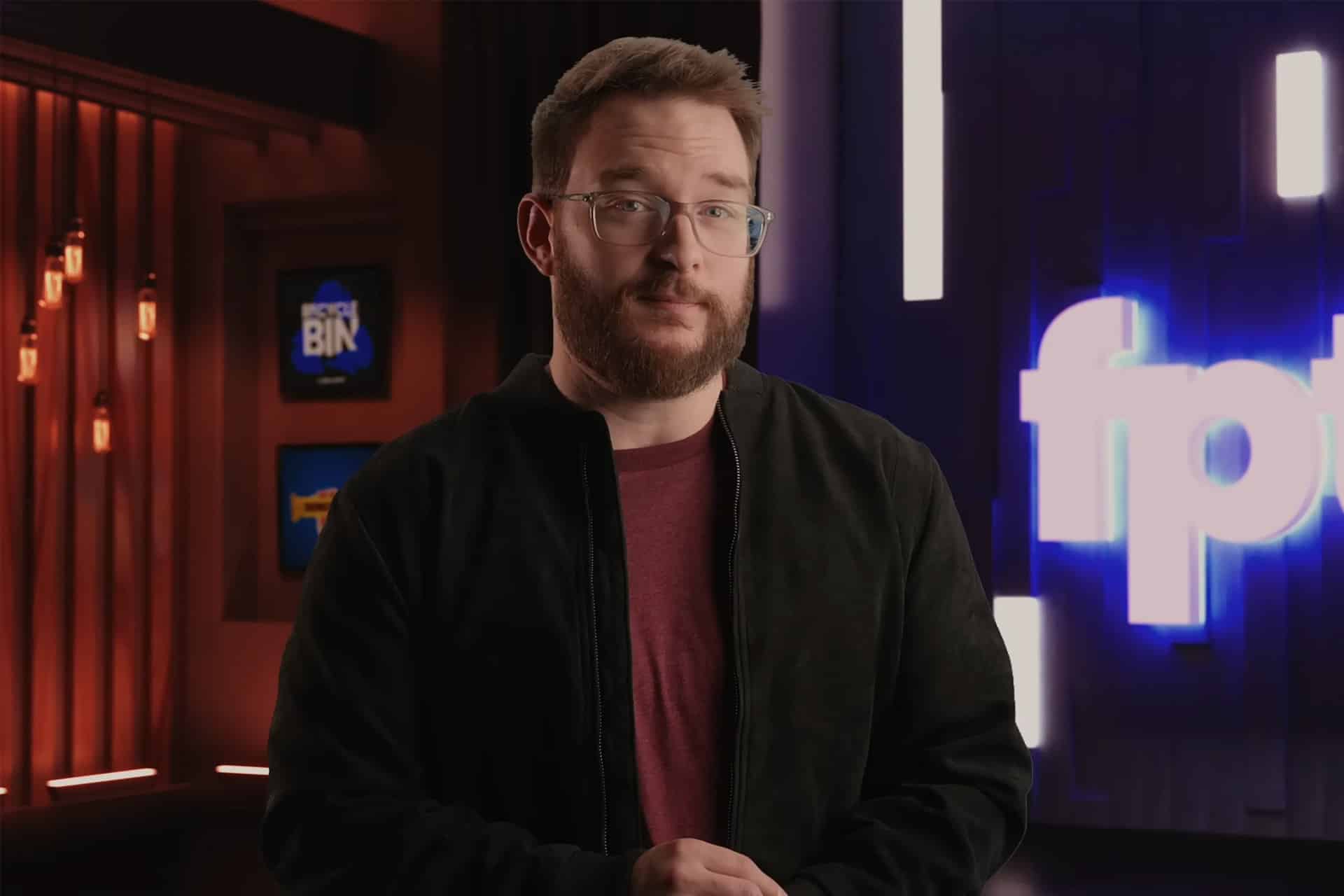 A man with short brown hair, glasses, and a beard stands indoors, wearing a maroon shirt and black jacket. Behind him are illuminated signs and a blurred blue logo—reminiscent of the backgrounds seen in Jon Prosser’s iOS 26 leaks videos.