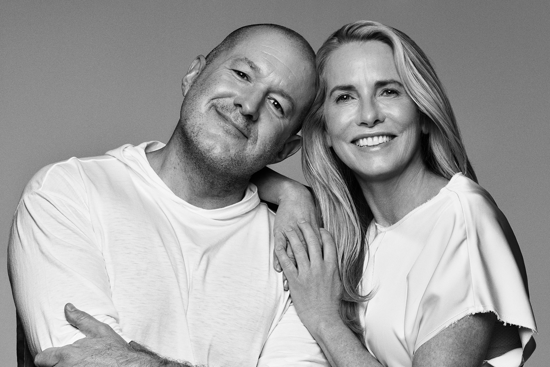 A smiling man and woman pose closely together against a plain background, both wearing light-colored clothing. The woman rests her hand on the man's shoulder, their relaxed demeanor reminiscent of Jony Ive's understated design philosophy.