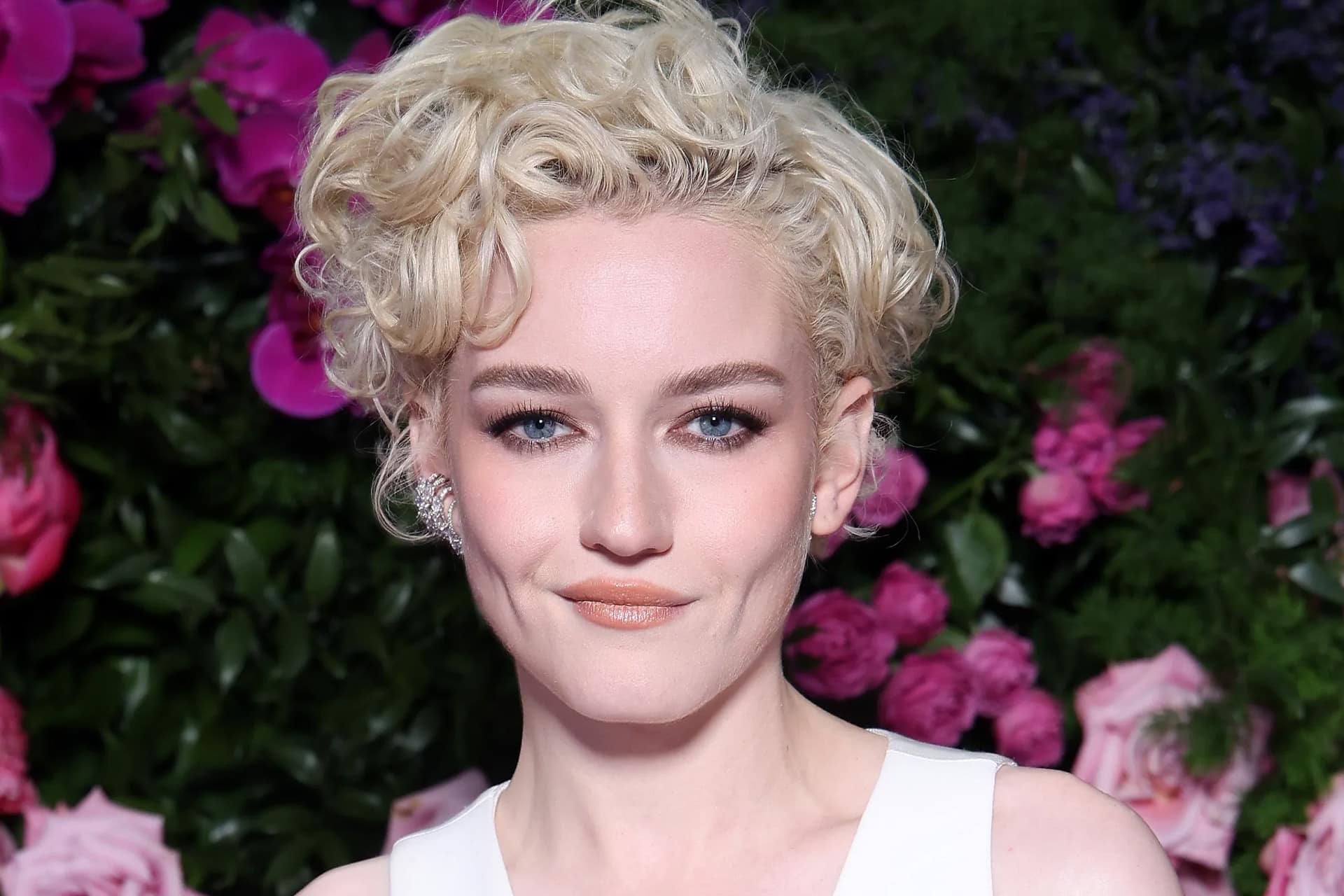 A person with short, curly blonde hair and light eyes poses in front of vibrant pink and purple flowers, channeling Julia Garner’s elegance in a white outfit and natural makeup.