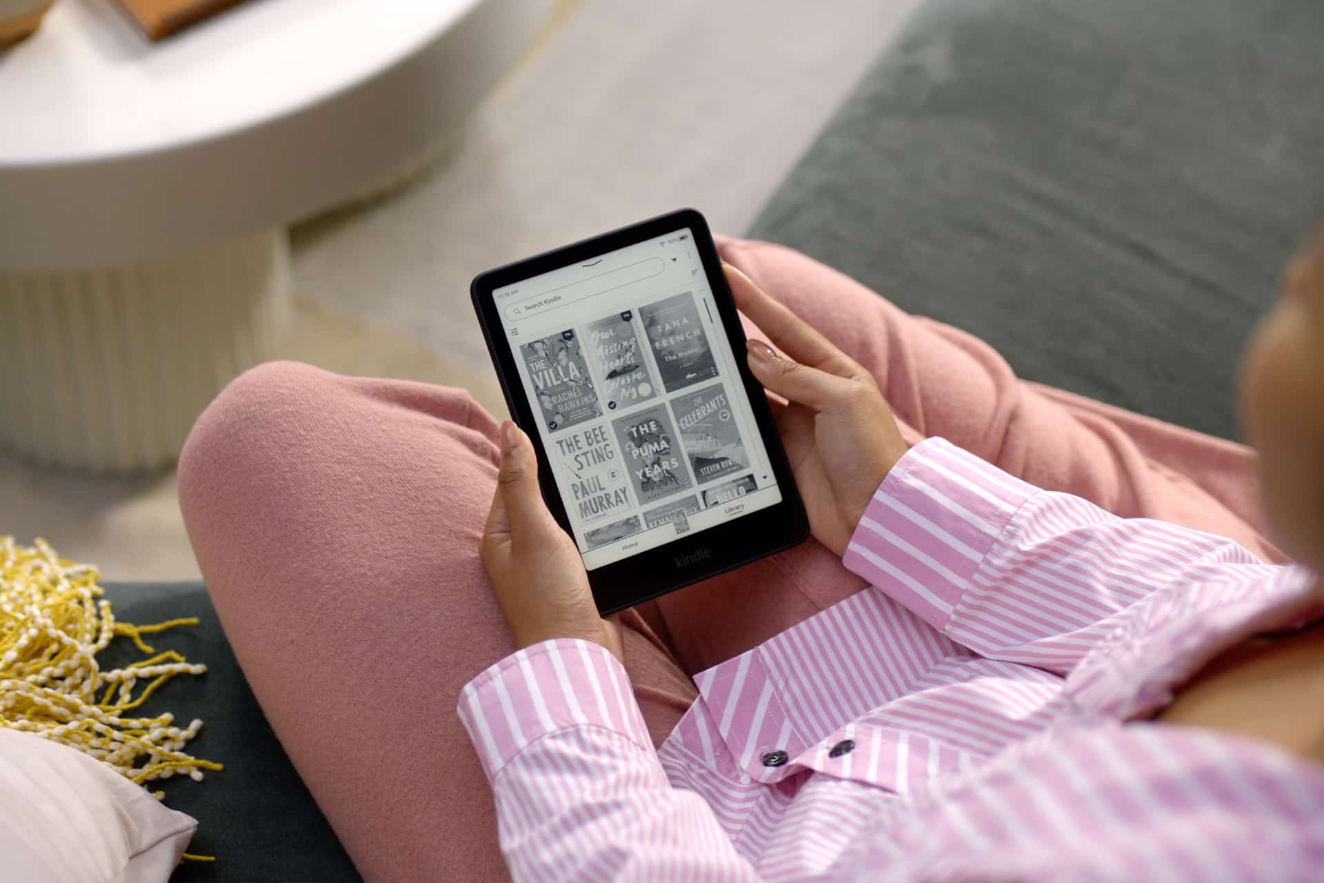 A person in pink pajamas sits on a couch holding an e-reader displaying book covers, some of which are suspected to be AI-generated books. A table with a yellow fringed item is visible nearby.