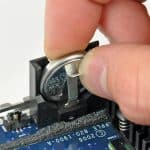 Mac PRAM - A hand is removing a round silver CMOS battery from a holder on a computer motherboard using a small metal tool to perform an NVRAM reset.