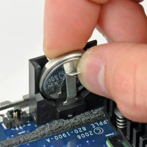 Mac PRAM - A hand is removing a round silver CMOS battery from a holder on a computer motherboard using a small metal tool to perform an NVRAM reset.