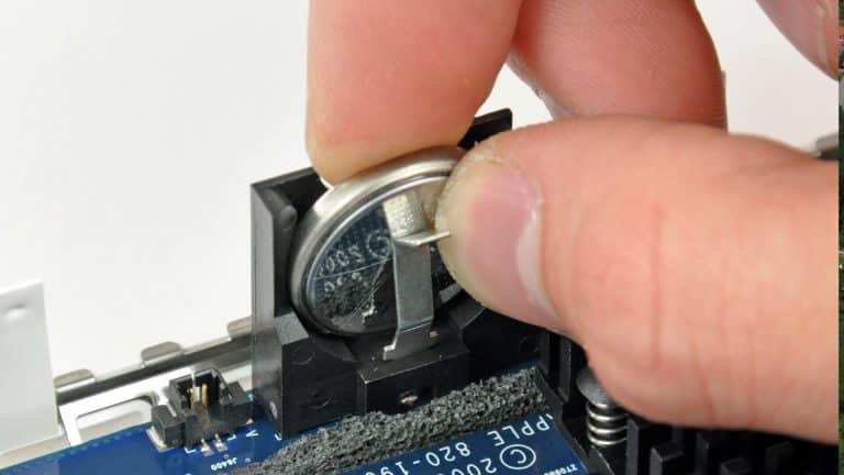 Mac PRAM - A hand is removing a round silver CMOS battery from a holder on a computer motherboard using a small metal tool to perform an NVRAM reset.