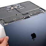 Hands are removing the bottom cover of a dark blue Apple laptop, revealing the internal components, including the battery and circuitry—a closer look that’s often necessary when addressing issues like MacBook Air Flexgate or troubleshooting display problems related to the MacBook Air screen.