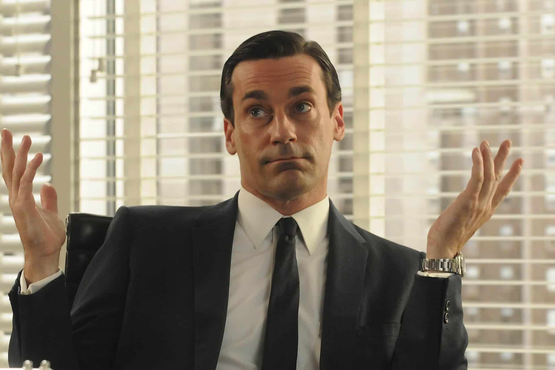 A man in a suit sits at a desk with his arms raised and palms up, appearing indifferent or confused—perhaps about his purchased TV shows—in an office with large windows and blinds in the background.