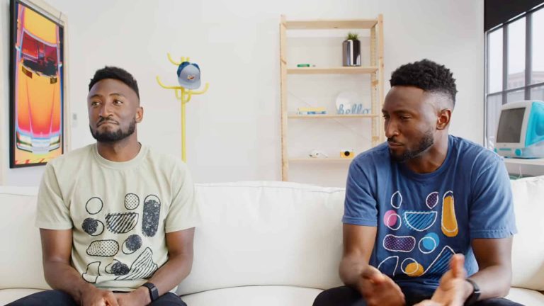 Two men who look identical sit on a white couch in a bright room; one looks thoughtful, the other gestures while speaking. Shelves, art, a coat rack, and a vintage computer are in the background—a subtle nod to the evolution from early tech to the era of Apple Intelligence on-device.