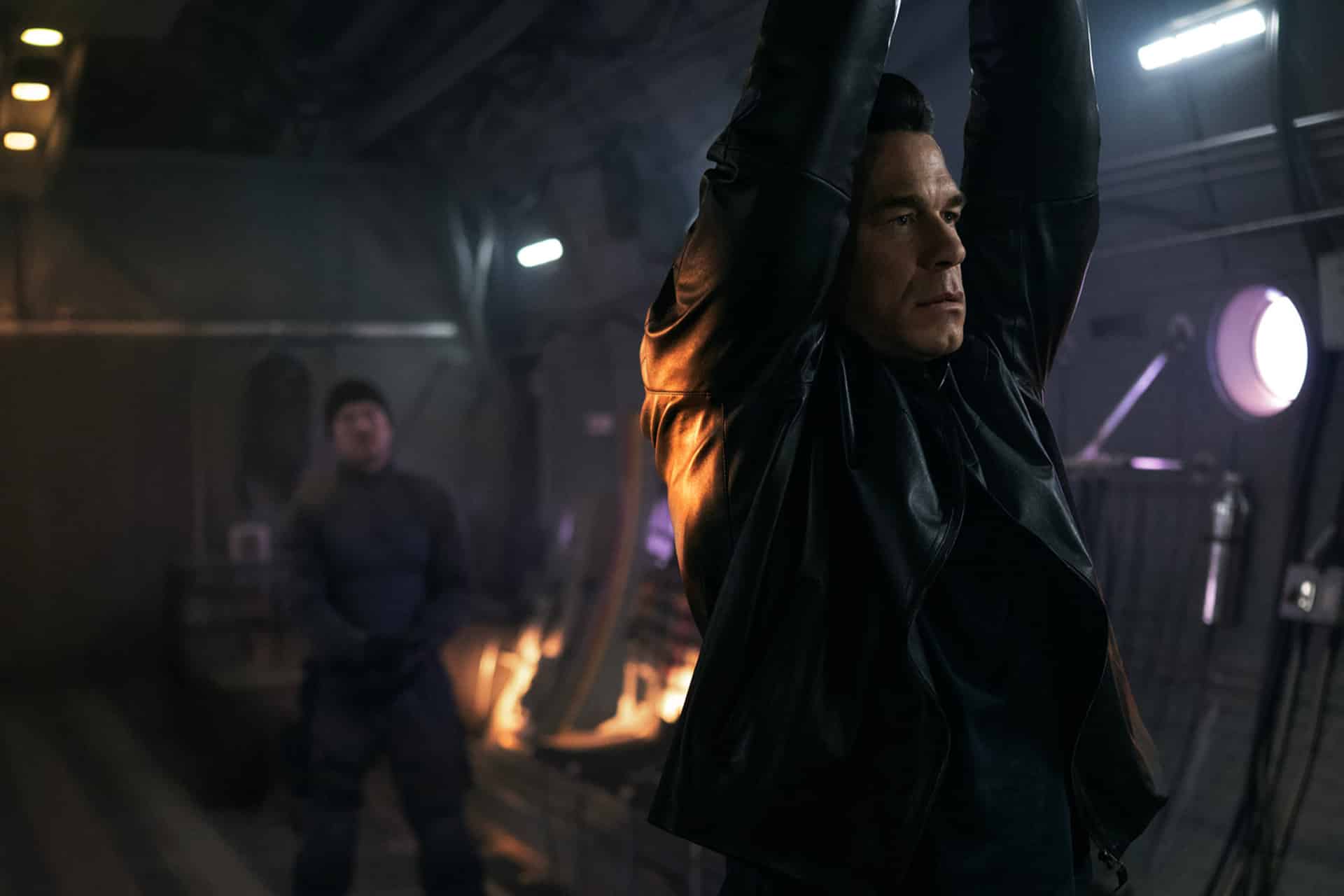 A man in a black leather jacket is hanging by his arms in a dimly lit room, while another person stands in the background watching him.