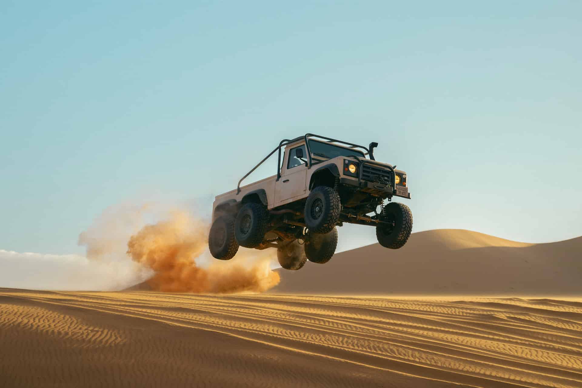 A white off-road vehicle jumps high above desert sand dunes, kicking up a cloud of dust under a clear sky.