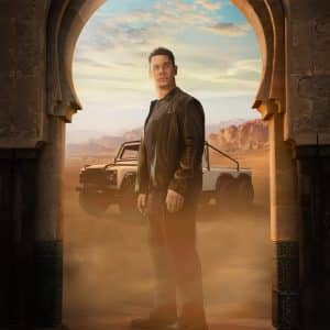 A man in a black leather jacket stands in an arched doorway with desert mountains and an off-road vehicle behind him, dust swirling around his feet.