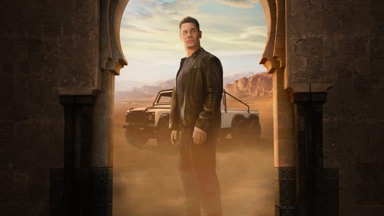 A man in a black leather jacket stands in an arched doorway with desert mountains and an off-road vehicle behind him, dust swirling around his feet.