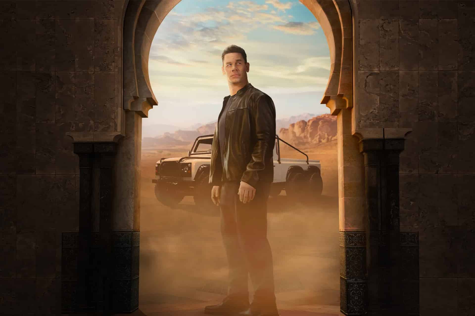 A man in a black leather jacket stands in an arched doorway with desert mountains and an off-road vehicle behind him, dust swirling around his feet.