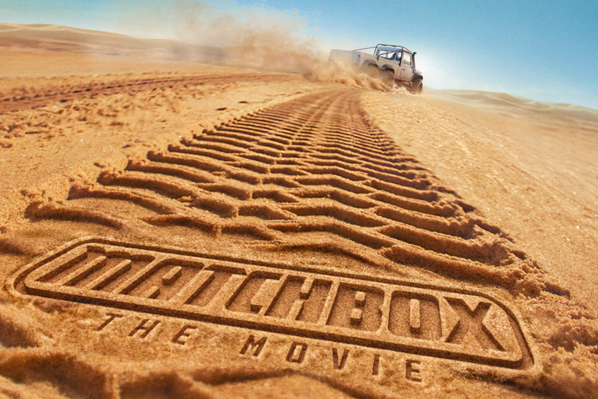 Matchbox The Movie - A vehicle drives across sandy dunes, leaving deep tire tracks. In the foreground, "MATCHBOX THE MOVIE" is carved into the sand, resembling a tire tread pattern. Dust rises behind the vehicle under a clear blue sky.