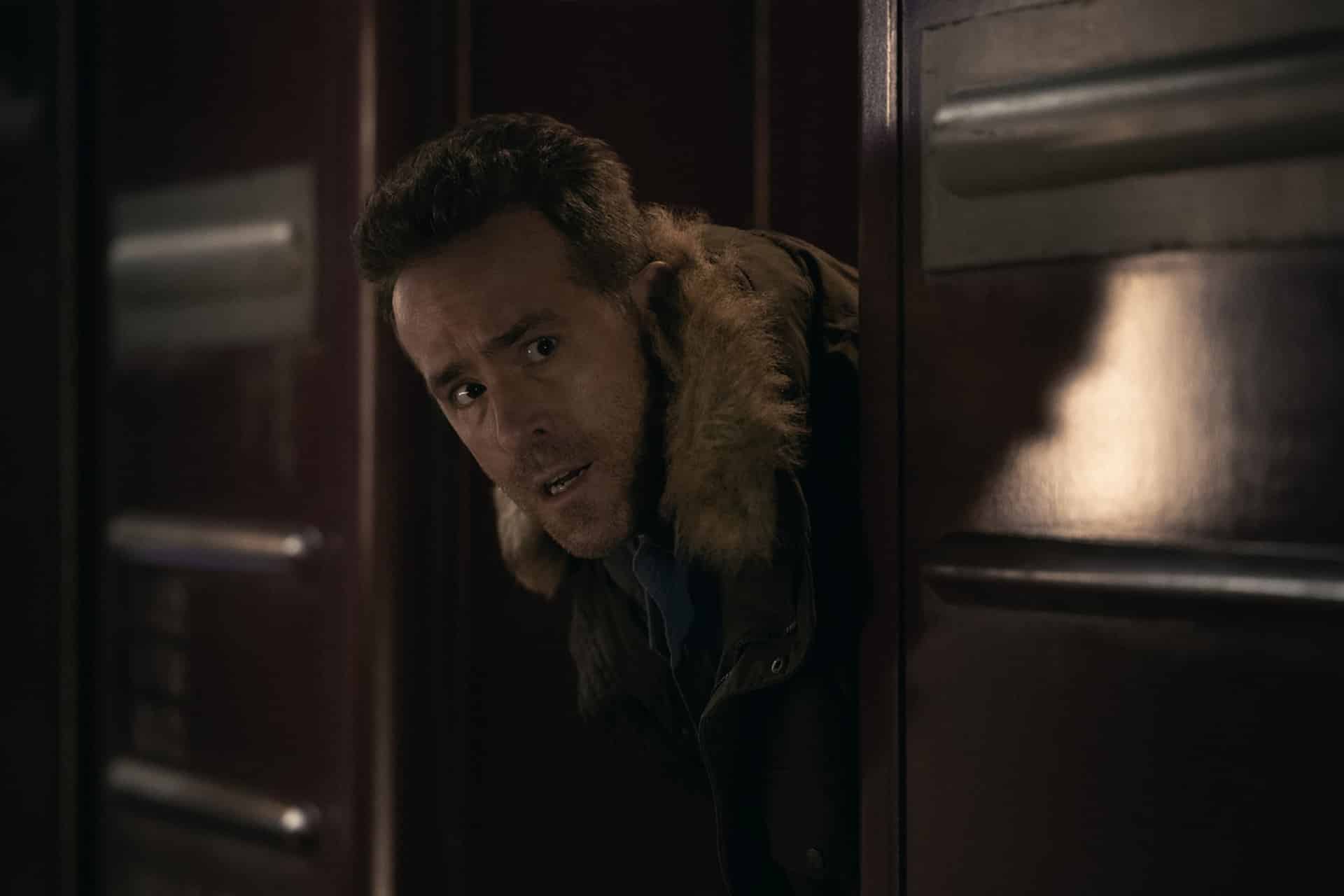 A man with short hair and a fur-lined jacket anxiously peeks out from behind a dark wooden door, with a worried expression on his face—a scene reminiscent of the suspense found in Apple Originals like Mayday on Apple TV.