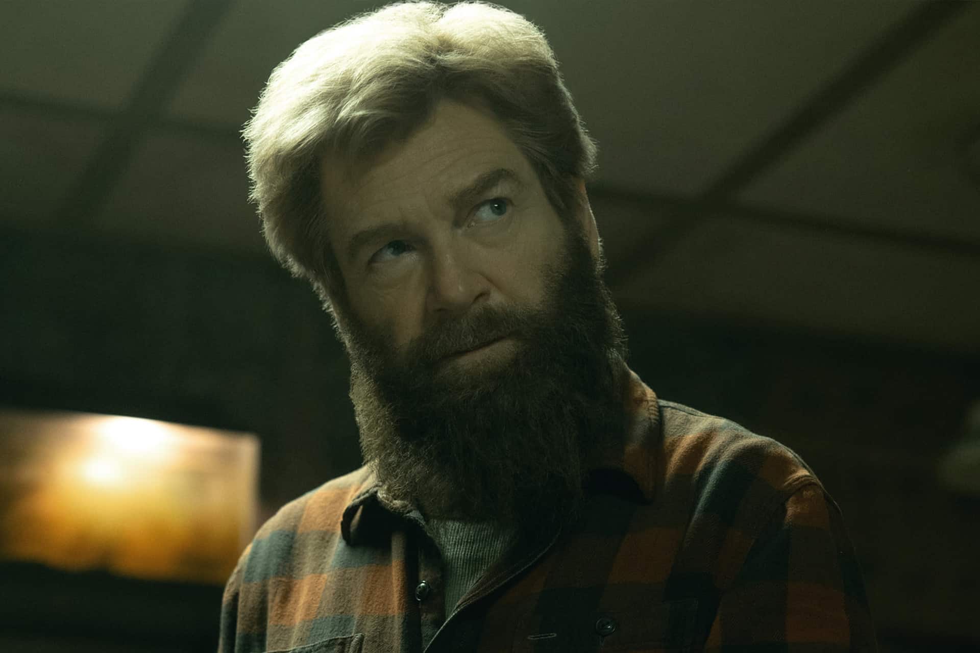 A man with a thick beard and tousled hair, wearing a plaid flannel shirt, looks upward with a thoughtful expression in a dimly lit room, reminiscent of the evocative storytelling found in Apple Original Films.