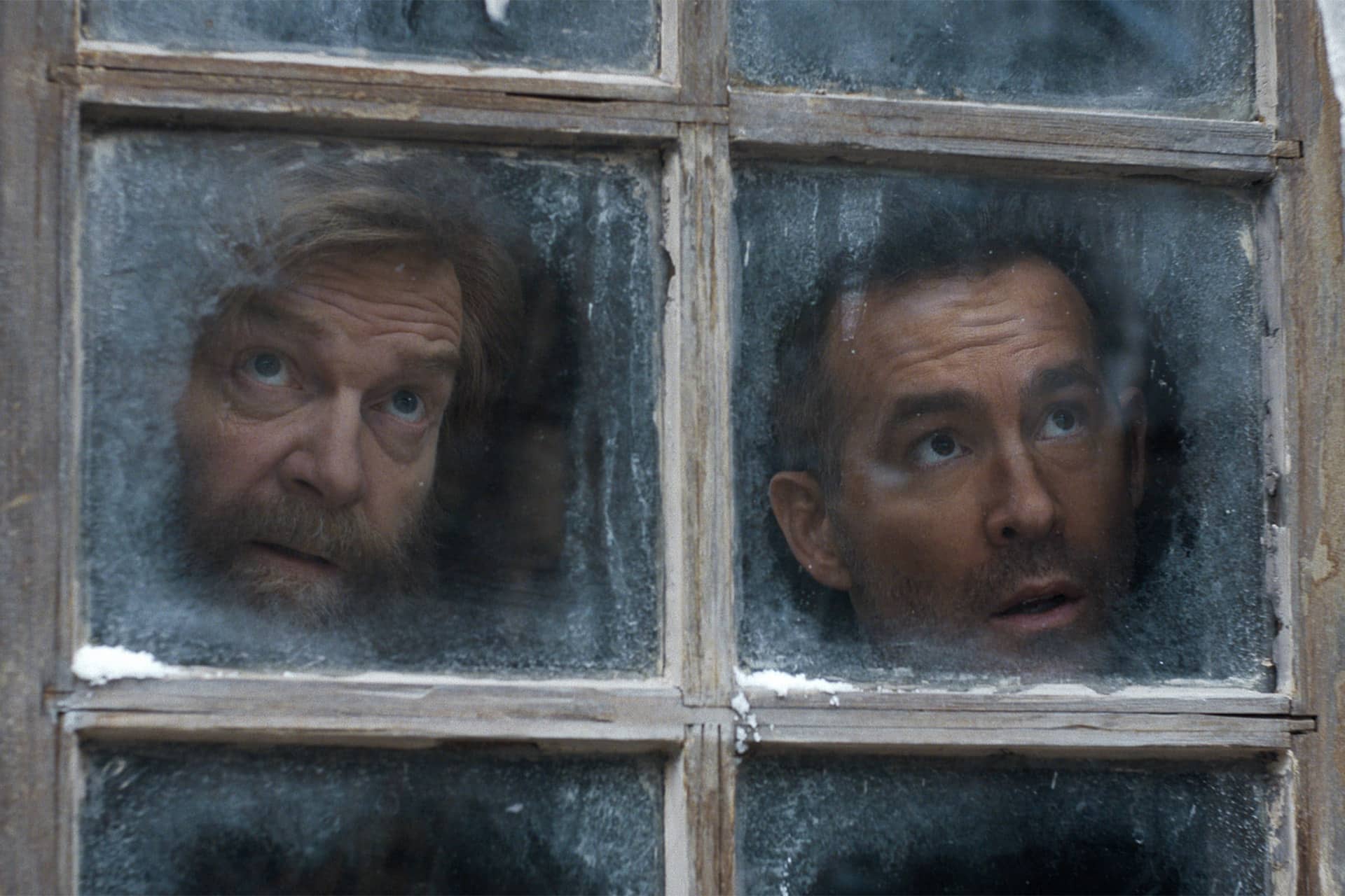 Two men with surprised expressions look through a frosty window from inside a building. Snow is visible on parts of the window frame, and the glass is partially covered with ice, creating a scene reminiscent of suspenseful moments often found in Apple Original Films.