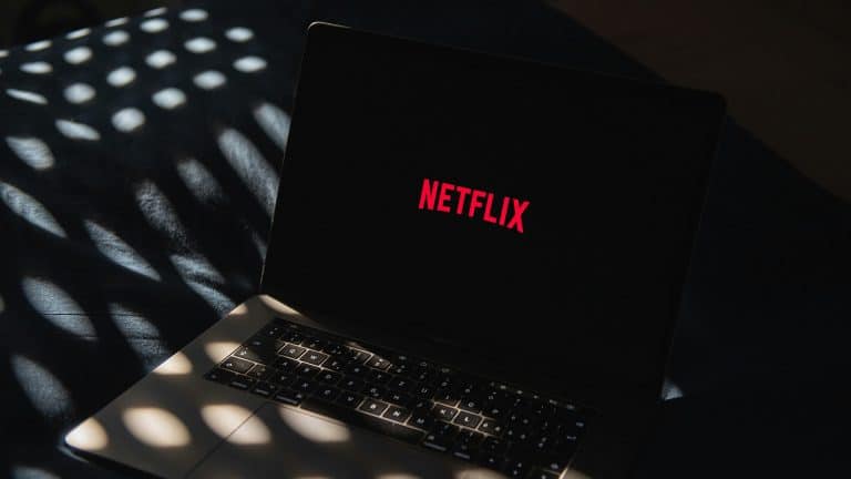 A laptop displaying the red Netflix logo on its screen sits on a dark surface, with patterned light spots shining across the keyboard and background—perfectly capturing the anticipation of diving into the latest streaming lineup.