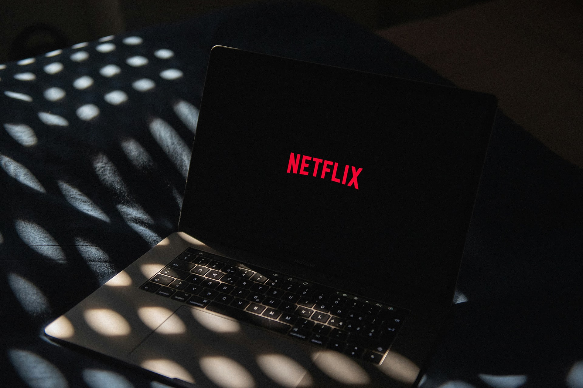 A laptop displaying the red Netflix logo on its screen sits on a dark surface, with patterned light spots shining across the keyboard and background—perfectly capturing the anticipation of diving into the latest streaming lineup.