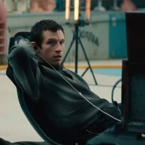 A person with short hair, wearing a textured shirt, leans back in a chair with one arm raised and earphones in, looking to the side. There is equipment and lights set up in the background of an indoor space, evoking the behind-the-scenes energy of a Neuromancer teaser for the upcoming Apple TV sci-fi Neuromancer series.