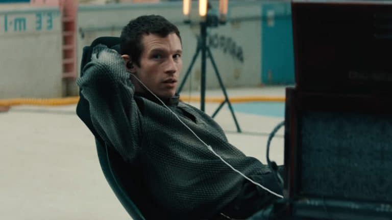 A person with short hair, wearing a textured shirt, leans back in a chair with one arm raised and earphones in, looking to the side. There is equipment and lights set up in the background of an indoor space, evoking the behind-the-scenes energy of a Neuromancer teaser for the upcoming Apple TV sci-fi Neuromancer series.