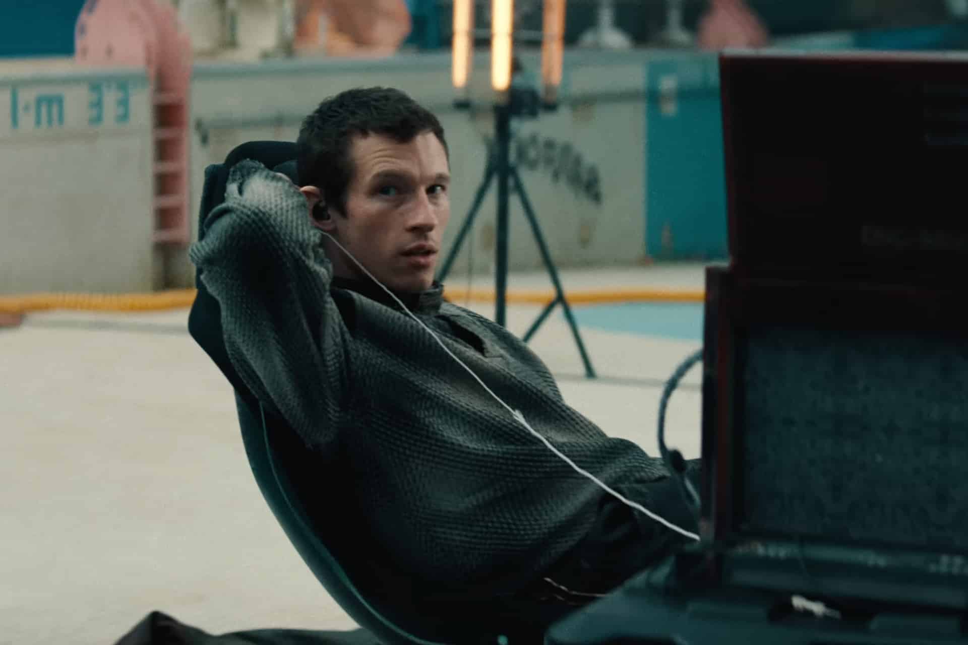 A person with short hair, wearing a textured shirt, leans back in a chair with one arm raised and earphones in, looking to the side. There is equipment and lights set up in the background of an indoor space, evoking the behind-the-scenes energy of a Neuromancer teaser for the upcoming Apple TV sci-fi Neuromancer series.