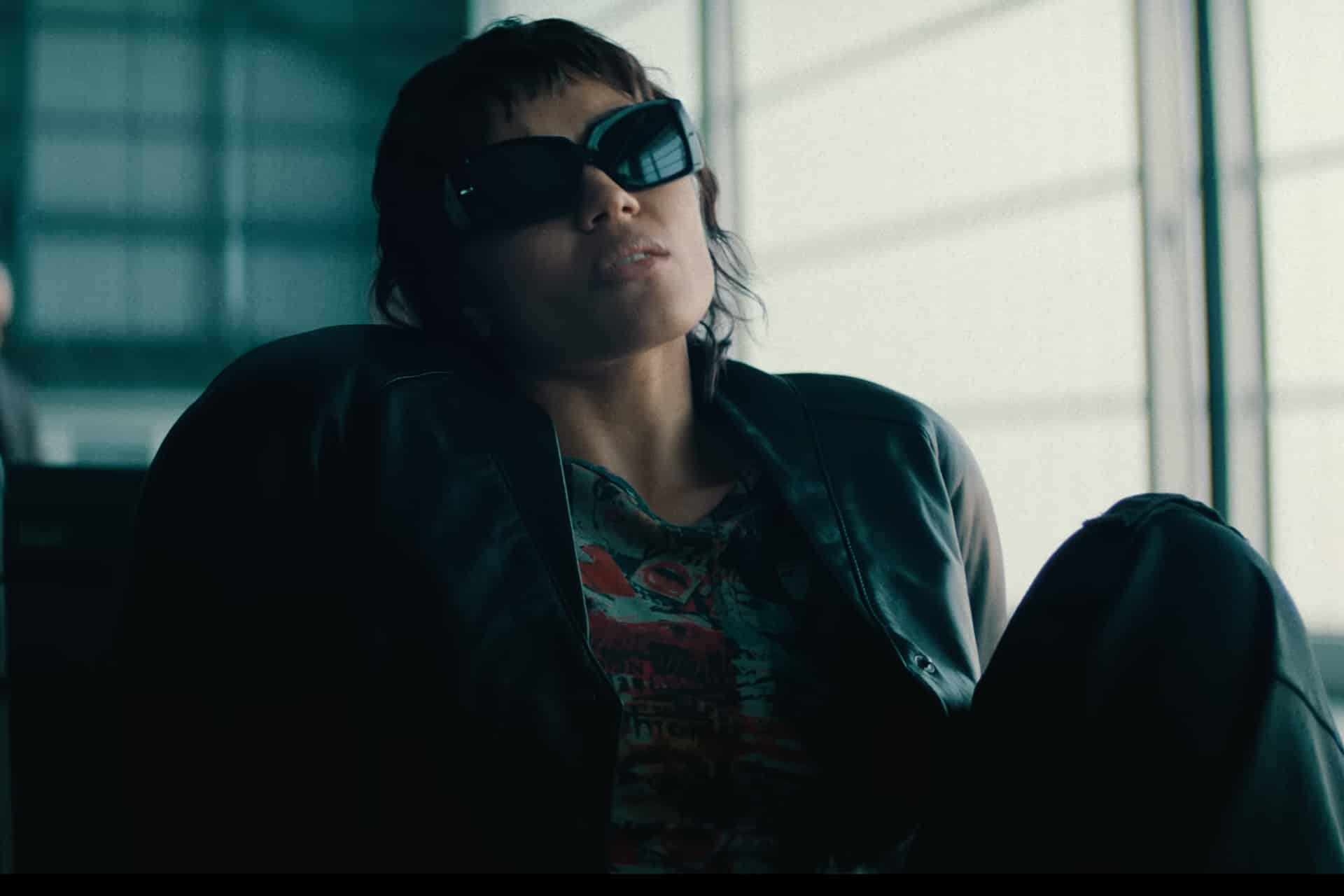 A person wearing dark sunglasses and a black jacket leans back in a chair, looking relaxed, with a window and modern building interior in the background—evoking the cool, cyberpunk vibe of the upcoming Apple TV sci-fi Neuromancer series.