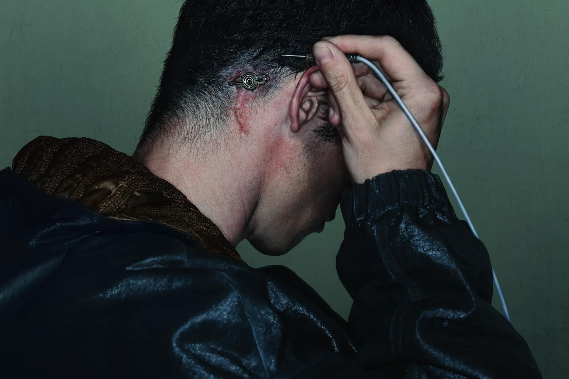 A person with short dark hair inserts a wire into a small port behind their ear, which is red and irritated, in this striking Neuromancer teaser. They wear a dark jacket and face slightly downward against a green background, echoing the cyberpunk atmosphere of the anticipated Apple TV sci-fi Neuromancer series.