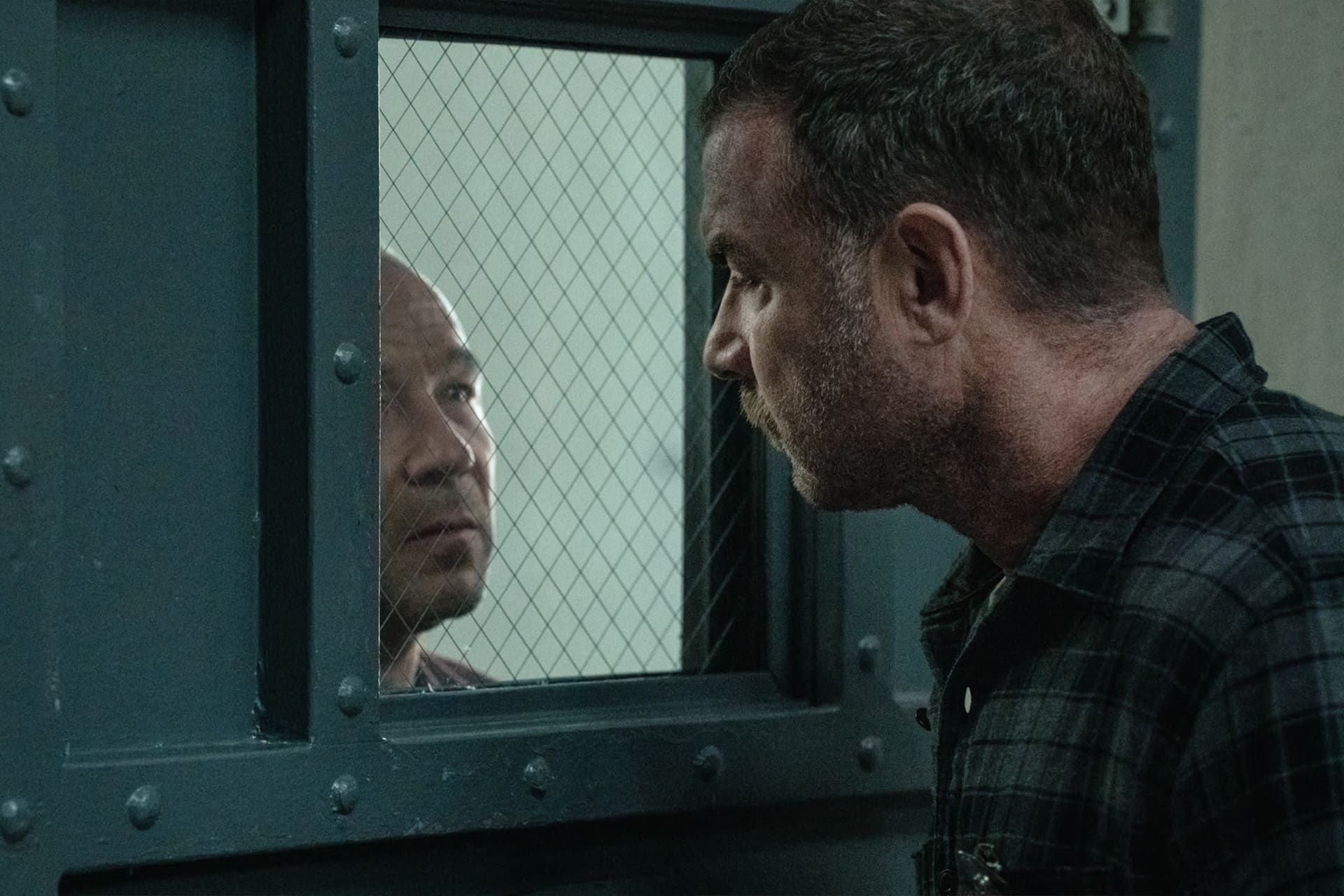 A man stands on one side of a metal door with a wired glass window, staring intently at another man—evoking the intense atmosphere of an Apple TV crime drama like Nocturne or a scene starring Liev Schreiber in a tense, prison-like setting.
