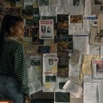 A woman in a striped sweater and jeans stands in front of a wall covered with maps, notes, photographs, and several missing person posters, looking concerned and deep in thought—a scene straight out of an Apple TV crime drama like Nocturne.