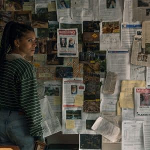 A woman in a striped sweater and jeans stands in front of a wall covered with maps, notes, photographs, and several missing person posters, looking concerned and deep in thought—a scene straight out of an Apple TV crime drama like Nocturne.