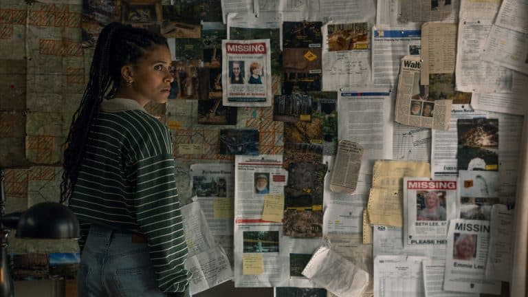 A woman in a striped sweater and jeans stands in front of a wall covered with maps, notes, photographs, and several missing person posters, looking concerned and deep in thought—a scene straight out of an Apple TV crime drama like Nocturne.