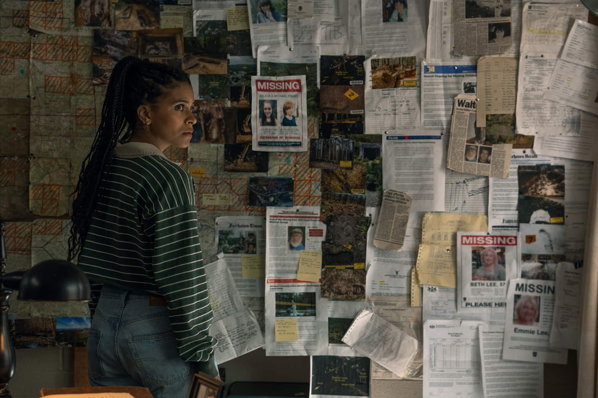 A woman in a striped sweater and jeans stands in front of a wall covered with maps, notes, photographs, and several missing person posters, looking concerned and deep in thought—a scene straight out of an Apple TV crime drama like Nocturne.