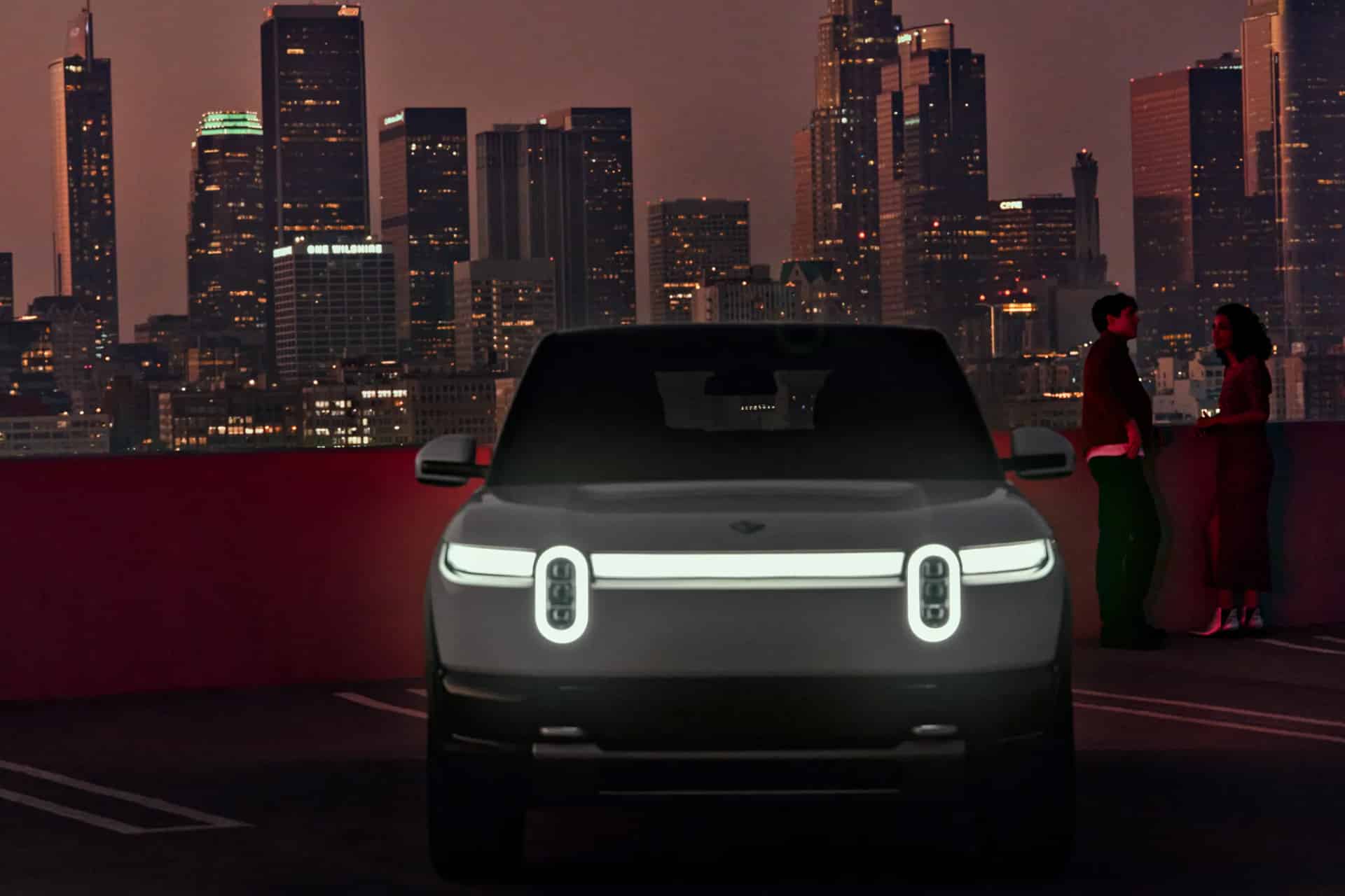 A white electric SUV, reminiscent of a Rivian Apple comparison, sits glowing on a rooftop at dusk. Illuminated headlights shine as two people stand by the railing, talking and enjoying the city skyline in the background.