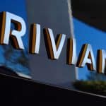 Close-up of the word "RIVIAN" in large, three-dimensional letters on a reflective, dark-colored surface, with outdoor scenery and perhaps a hint of RJ Scaringe’s innovative spirit faintly mirrored.