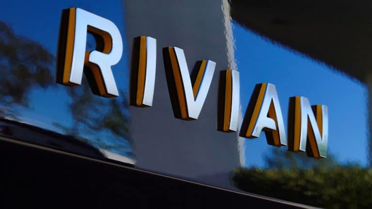 Close-up of the word "RIVIAN" in large, three-dimensional letters on a reflective, dark-colored surface, with outdoor scenery and perhaps a hint of RJ Scaringe’s innovative spirit faintly mirrored.