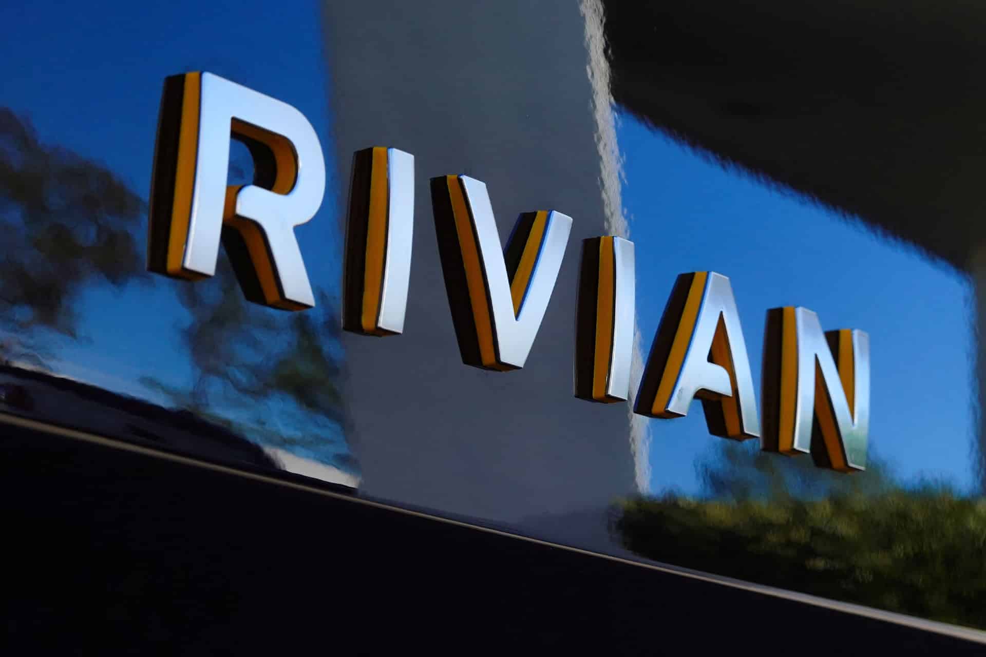 Close-up of the word "RIVIAN" in large, three-dimensional letters on a reflective, dark-colored surface, with outdoor scenery and perhaps a hint of RJ Scaringe’s innovative spirit faintly mirrored.