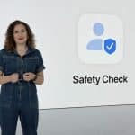 A woman in a denim jumpsuit stands next to a large screen displaying an icon of a person with a checkmark shield and the words "Safety Check" on a white background, emphasizing features like shared access and Find My for enhanced security.