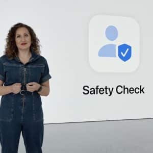 A woman in a denim jumpsuit stands next to a large screen displaying an icon of a person with a checkmark shield and the words "Safety Check" on a white background, emphasizing features like shared access and Find My for enhanced security.