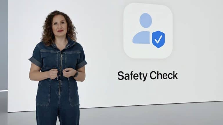 A woman in a denim jumpsuit stands next to a large screen displaying an icon of a person with a checkmark shield and the words "Safety Check" on a white background, emphasizing features like shared access and Find My for enhanced security.