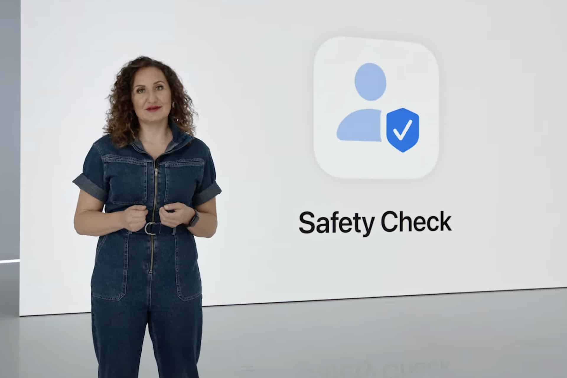 A woman in a denim jumpsuit stands next to a large screen displaying an icon of a person with a checkmark shield and the words "Safety Check" on a white background, emphasizing features like shared access and Find My for enhanced security.
