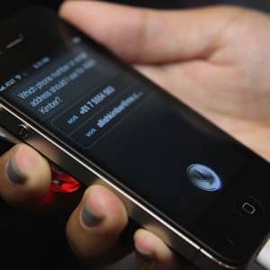 A hand holds a black iPhone displaying Siri, with a contact request on the screen—showcasing Siri’s integration with Wolfram Alpha. The phone is plugged into a white charging cable.