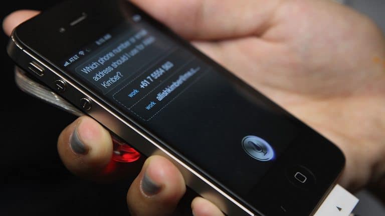 A hand holds a black iPhone displaying Siri, with a contact request on the screen—showcasing Siri’s integration with Wolfram Alpha. The phone is plugged into a white charging cable.