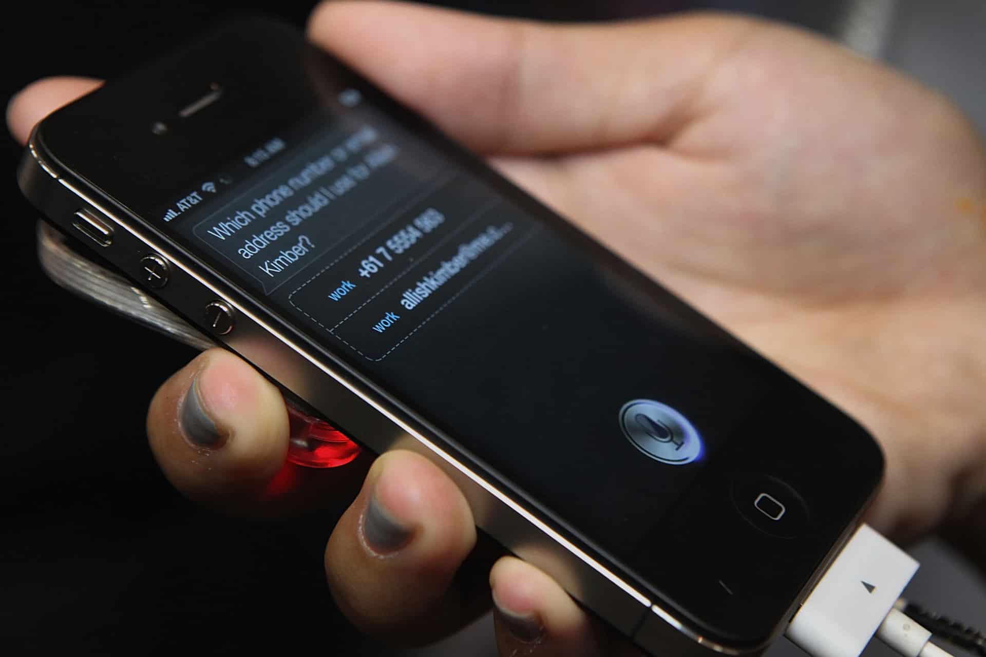 A hand holds a black iPhone displaying Siri, with a contact request on the screen—showcasing Siri’s integration with Wolfram Alpha. The phone is plugged into a white charging cable.