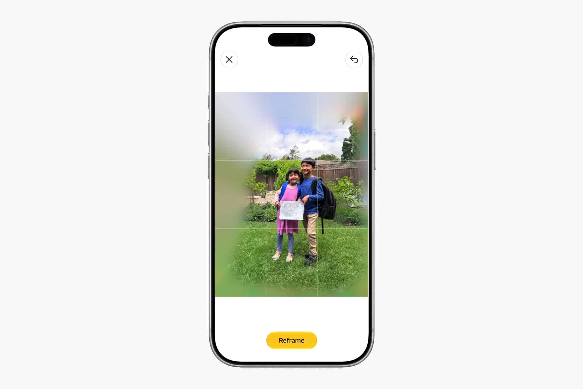 A smartphone screen shows a photo of two students posing outdoors in a garden, with a digital blur effect around them and a “Reframe” button at the bottom, highlighting advanced photo editing features in the Photos app to enhance your spatial memories.