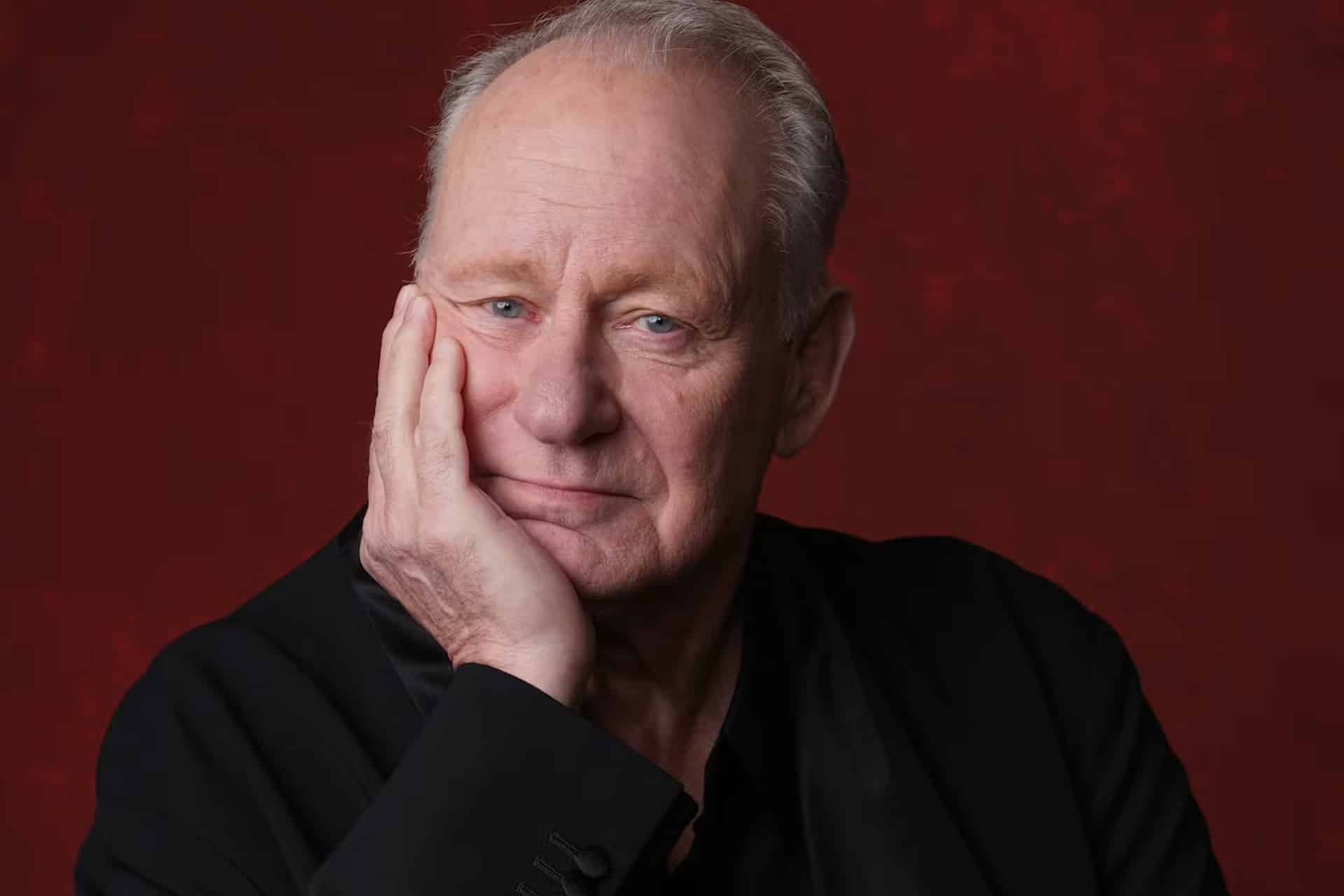 An older man with short gray hair and a thoughtful expression rests his face on his hand. He wears a black jacket and sits in front of a deep red background, evoking the contemplative mood of Homeland, produced by Alex Cary for Sony Pictures Television.