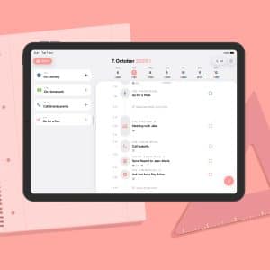 A tablet displays a digital planner for October 7, 2025, showcasing tasks and a calendar—perfect for digital planning enthusiasts. It rests on a spiral notebook and a pink triangular ruler, set against a pink background.